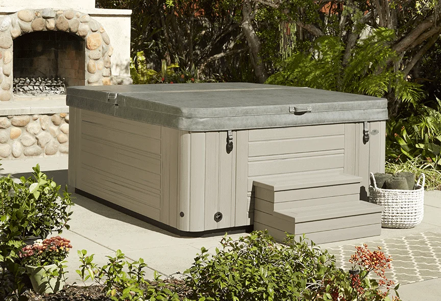 Installed hot tub with gray cover, tan siding, and built-in steps in landscaped backyard