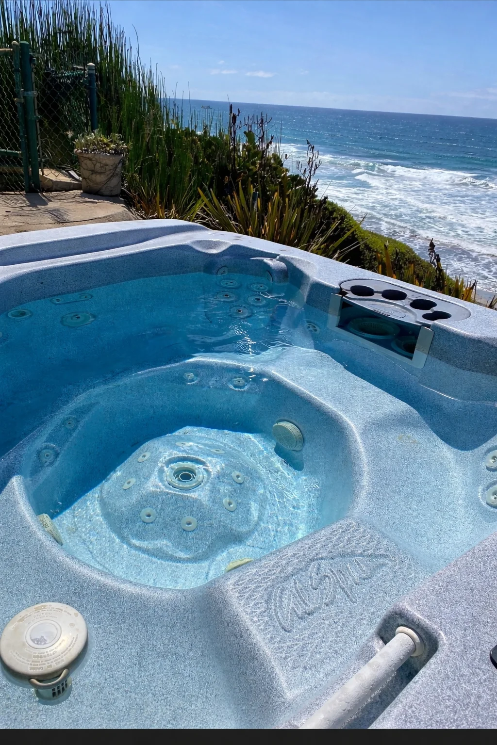 Blue hot tub with dual seats, jet nozzles, and control panel on oceanfront deck with coastal plants