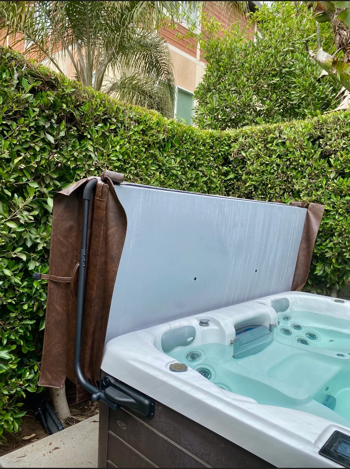 Hot tub with aluminum privacy screen frame, gray composite panel, and brown suede cover among green hedges